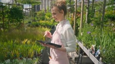 Çağdaş serada yetiştirme sistemini kontrol eden tablet bilgisayar kullanan pozitif kadın bahçıvan. Sera işçisi bir tablet bilgisayar kullanarak bitkilerin büyümesini kontrol ediyor.