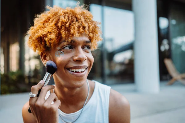 Positive multiracial charismatic boy preparing makeup at his face with visage brush while standing at the street and looking at the camera. Self identify and freedom concept