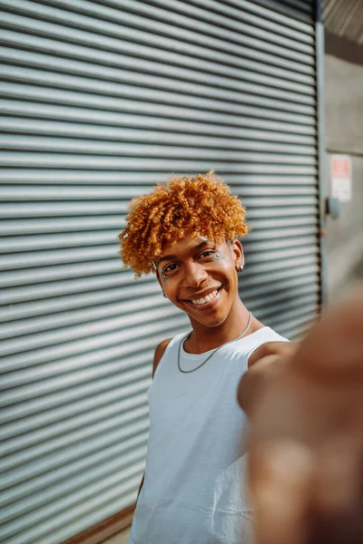 Vertical view of the teenager multiracial man smiling confident while making selfie by the camera at the street. Guy with makeup using smartphone camera outdoors in city street. People and photography