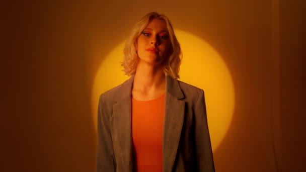 オレンジ色のスタジオの背景の上にポーズをとるジャケットの若いエレガントな女性 ファッションスタジオのショットコンセプト 肖像画 — ストック動画