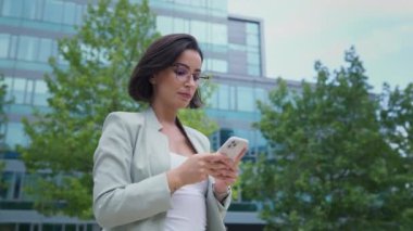 Serious businesswoman standing near office modern business center and writing message at her smartphone. Lady boss waiting for colleague 