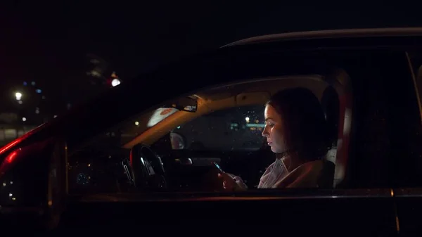 夜は車の中でスマホを使う美人ビジネスウーマン 運転席の携帯電話で働く忙しい女性 バックにボケとネオンライト 画面デバイスによって照明された — ストック写真