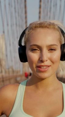 Kulaklıkla müzik dinleyen ve köprüdeki kameraya bakan sarışın seksi sporcunun portresinin dikey görüntüsü. Koşu için hazırlanan bayan koşucu