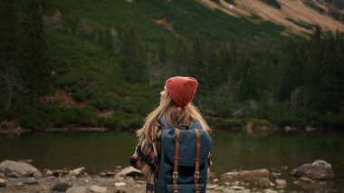 Back view of stylish hipster woman holding backpack and looking at mountain view while relaxing in nature. Travel and wanderlust concept. Amazing chill moment