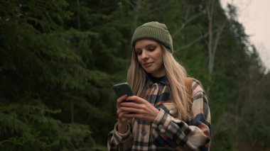 Girl tourist walking in forest and using smartphone gps. Woman hiking and typing message on smartphone. Female hiker preparing taking smartphone pic of mountains and forest