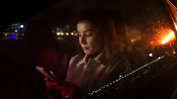 严肃的女商人在开车时使用智能手机 晚上在豪华轿车上下班时坐在那里 城市街道上有工作霓虹灯标志 — 图库照片