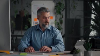 Office work. Senior businessman in casual shirt looking at the tablet screen while sitting at the table. Mature man preparing to the morning meeting