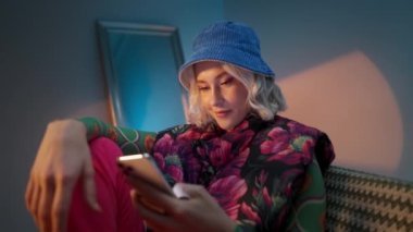 Moda kadını koltukta oturuyor, telefona gülümsüyor ve akşam stüdyo arka planında akıllı telefondan internete bakıyor.