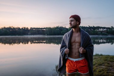 Winter swimming. Serious guy with towel at his shoulders walking at the forest near the water and preparing for the swimming in the cold winter lake at morning