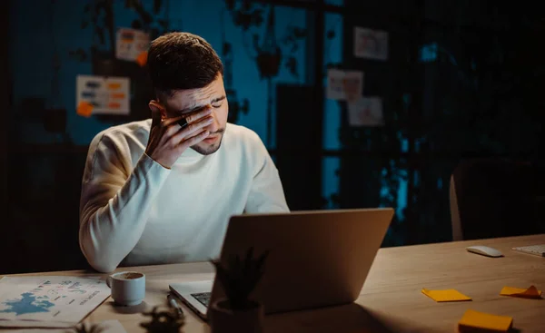 Tired man wearing stylish casual clothes rubbing eyes while working laptop and trying hurry up to deadline. Sad guy sitting in the modern office space. Office employee at evening