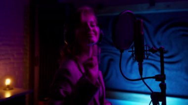 Funky singer woman using microphone and headphones, while recording song in neon light studio. Creative hobby and dream to be star