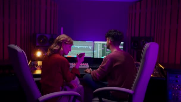 在音乐录音室与男性音频工程师一起工作的同时 拥有智能手机和听歌曲的创意女性 两位同事用混合程序软件创作了很酷的歌曲 创意艺术家 — 图库视频影像