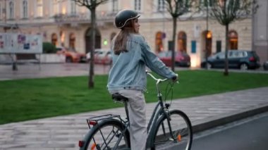 Yağmurlu bir akşam şehrinde bisiklet süren kasklı kızın arka görüntüsü. Çevre dostu ofisinin keyfini çıkaran genç bir kadın.