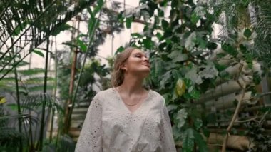 Arka planında yeşil yapraklar ve beyaz camlarla botanik bahçesinde bir kız. Mutlu kadın temiz havanın ve ormanların tadını çıkarıyor.