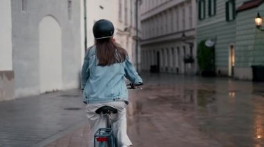 Şehir merkezinde yağmurlu cadde boyunca bisiklet süren güzel genç bir kadının arka manzarası. Etkin yaşam tarzı kavramı