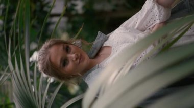 Bahçede poz veren güzel sarışın kadının dikey portresi. Rahat genç kız tropikal ağaçların etrafında dikilip ormanların tadını çıkarıyor.