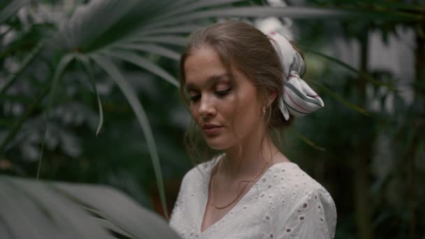 オレンジ色の植物の間に立っている白いドレスを着ている若い女性の肖像画 温室や植物園でポーズするファッショナブルな少女 — ストック動画