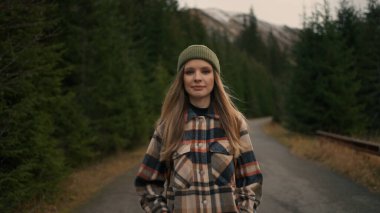 Sarışın kadın gezgin, doğa ile çevrili dağlarda yolda yürürken kameraya bakıyor. Seyahat gezisi seyahat tutkusu konsepti