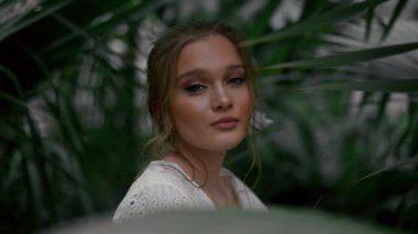 Açık makyajlı, bahçe resminde poz veren çarpıcı sarışın kadın. Rahat genç kız tropikal ağaçların etrafında dikilip ormanların tadını çıkarıyor.