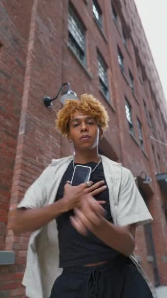 赤い都市の壁の背景に彼の手にスマートフォンで曲がった髪の歌と踊る楽しい10代の男の垂直な景色 ストリートライフスタイル — ストック動画
