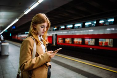 Genç bir kadın metro istasyonunda duruyor ve cep telefonuna bakıyor. Genç bir kadın cep telefonu başvurusu ile metro bileti alıyor.