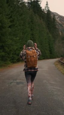Back view of the young woman traveller walking in middle of empty road in mountains, carrying backpack, enjoying of the nature. Travel road trip wanderlust concept