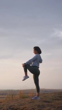 Caucasian female runner standing and warming up in preparation for a jogging run. She is located on a high cliff with beautiful mountains in the background