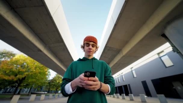 Uśmiechnięty Nastolatek Hipster Smsuje Smartfona Przy Nowoczesnym Zbliżeniu Miasta Stylowy — Wideo stockowe
