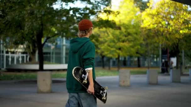トレンディなクールな男はスケートボードを保持し 都市公園を散歩しながら離れて見ています 人々のライフスタイルコンセプト — ストック動画