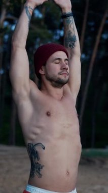 Young half naked sportsman warming up and training in pine forest during the winter. Man stretches muscles before winter swimming