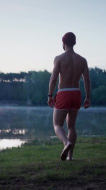 Back view of the good looking man in swim shorts going to the lake through the grass and looking at the water while preparing for the swimming in the winter morning. Ice swimming concept