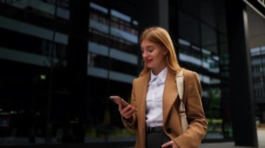 Neşeli kadın menajer modern akıllı telefon bulutlu bir günde şehirde yürüyor. Hareketli mesajları okuyan çekici bir iş kadını. Mutlu kadın CEO şık beyaz gömlek giyiyor telefon ekranı gibi 