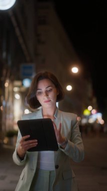 Vertical view of the beautiful young woman using tablet computer while walking through night city street full of lights. Portrait of gorgeous smiling female using her device