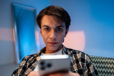 Handsome trendy young model posing at the evening studio background, sitting in a armchair and browsing on his mobile phone