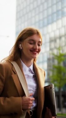 Kulaklıklı mutlu iş kadınının şehir merkezine giderken laptopuyla müşteriyle konuşurken dikey görüntüsü.