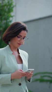 Active positive lady boss walking through the street and messaging at her phone with colleagues. Confident woman manager in formal wear taking a break and using mobile