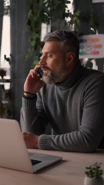 彼のオフィスで働く大人の男性は 顧客や同僚とのコミュニケーションのためにスマートフォンやラップトップを使用しています コンピュータ画面で見ている携帯電話を話す深刻なシニア男 — ストック動画