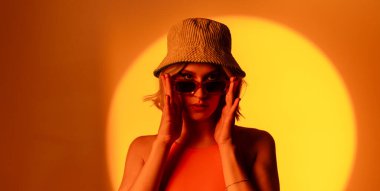 Fashionable young model wearing trendy sunglasses and stylish hat against orange background posing infront of camera in studio