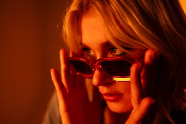 Stylish female model with light natural makeup wearing trendy sunglasses against orange background. Glamorous woman posing in studio