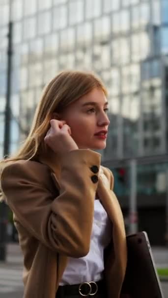 ハッピービジネスウーマンがイヤホンを挿入し 市内中心部に沿って彼女のラップトップと一緒に歩いている間にクライアントに電話するという垂直な見解 オフィスへの彼女の方法 — ストック動画