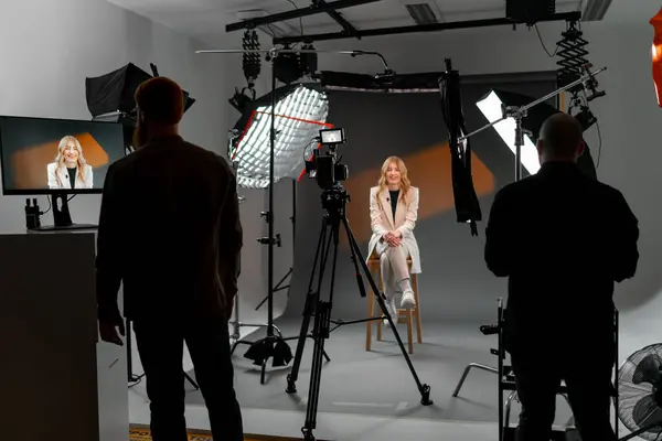 A woman is seated on a stool in a studio, facing a camera. The setting is an art event with visual arts in the background, creating a fun and entertaining atmosphere.