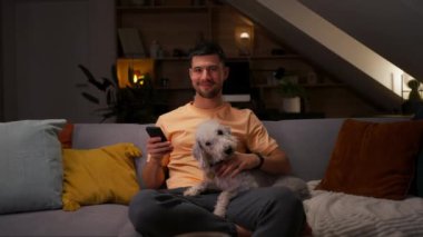 Genç adam elinde cep telefonu, kameraya bakıyor ve sevimli köpeğini kucaklıyor ve akşam odasında poz veriyor.
