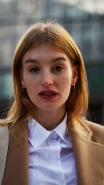 Genç bayan ofis müdürünün dikey görüntüsü bulutlu gökyüzünün önünde dikey duruyor ve gülümsüyor. Kendine güvenen stil sahibi iş kadını dışarıda rahatlıyor. Ofiste poz veren güzel bayan.