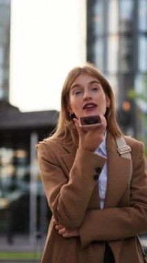 Şehirde cep telefonu kullanan ceketli, gülümseyen bir iş kadınının dikey görüntüsü.