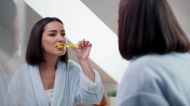 Mutlu bir kadın, bir elinde diş fırçasını tutarken büyük bir gülümsemeyle aynanın karşısında dişlerini fırçalıyor. Özel bir etkinliğe hazırlanıyor ve hijyen jestini paylaşıyor.