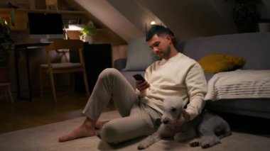 Halıda köpekle otururken cep telefonu tutan ve evcil hayvanını okşayan genç bir adam. Erkek ev sahibinin portresi akşamları evde oturup hayvanları tırmalıyor.