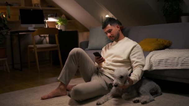 Joven Sosteniendo Teléfono Móvil Mientras Está Sentado Alfombra Con Perro — Vídeo de stock