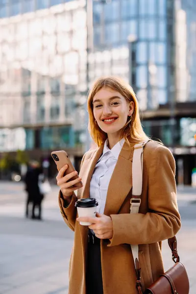 顔に笑顔の女性がコーヒーと携帯電話のカップを持っており レザージャケットの袖を持つ電気ブルーのブレーザーを着ています 彼女は特別なイベントに旅行する準備をしている間 ジェスチャー ストック写真