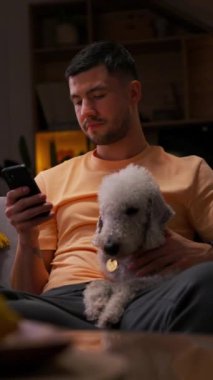 Yüz kılları ve şapkası olan bir adam koltukta oturuyor, kollarında küçük beyaz bir köpek tutuyor, telefonuna bakıyor.