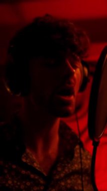 Singer man holding hands on headphones, while recording song in red light studio. Creative hobby concept
